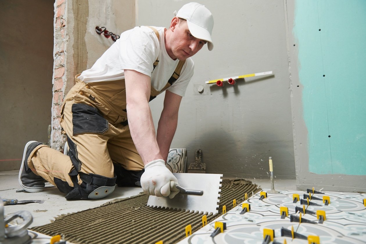 Professional tiler placing ceramic tiles on a bathroom floor indoors during a home renovation, with tools and materials visible.