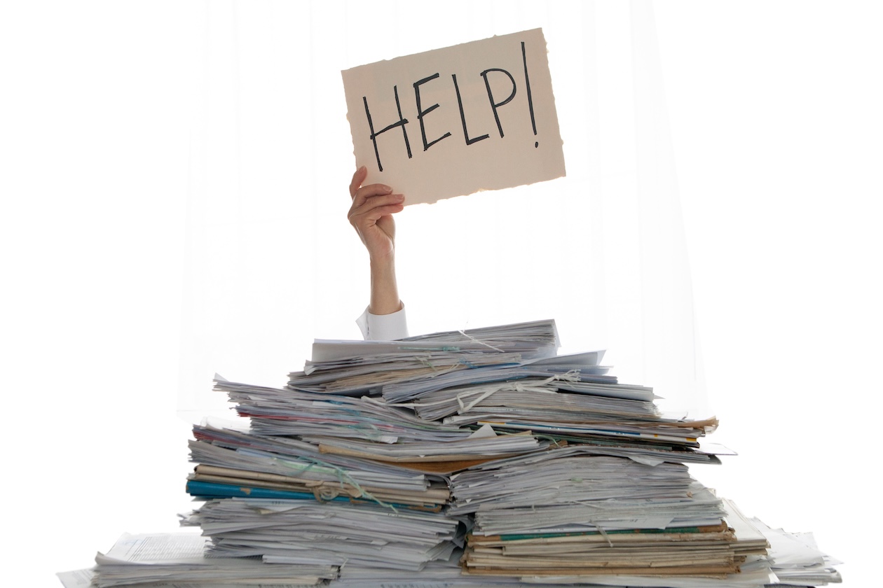 Overwhelmed office worker buried under a pile of paperwork holding a help sign, symbolizing inefficiency without flooring inventory software.