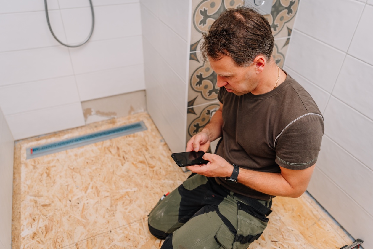 Flooring installer using a smartphone to manage job details in a renovated bathroom, representing cloud-based flooring software.